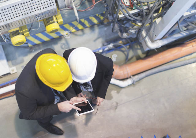 Two men in hard hats on a factory floor consulting over a tablet.