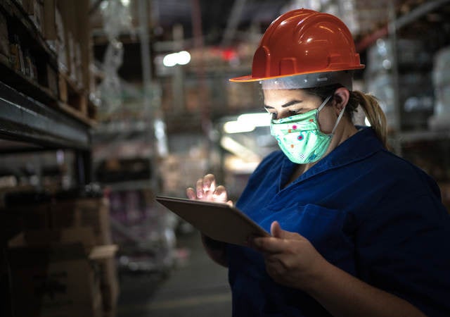 Portrait of mid adult woman wearing face mask using digital tablet - working at warehouse/industry.