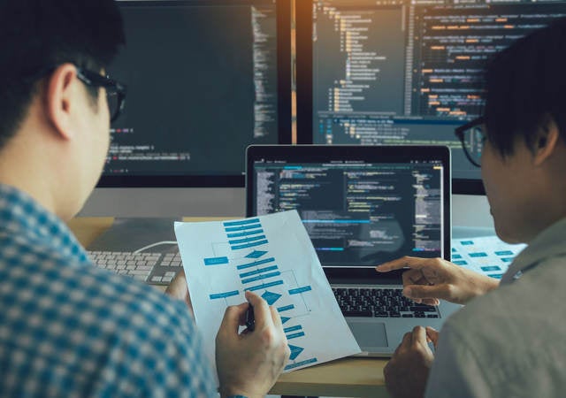 Two engineers examining a process flow on paper with coding on computer screens in the background.