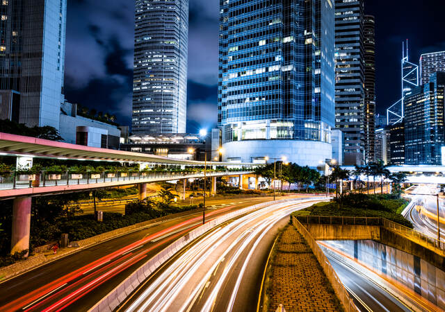 A multi-lane highway is brightly illuminated by roadway lighting, with a modern cityscape on the horizon.