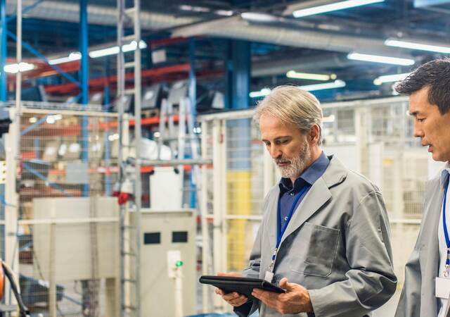 Two engineers walking around in a factory with a digital tablet and talking. Robotic arm in the background.