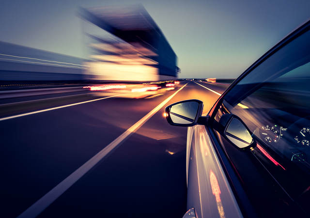 Silver car with side-view mirror driving on highway beside heavy duty truck at night