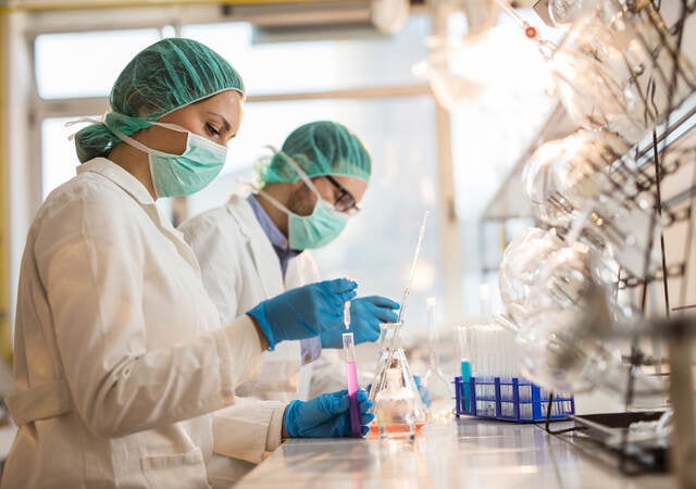 Two scientists wearing facemasks and examining chemical substances in test tubes in a laboratory.