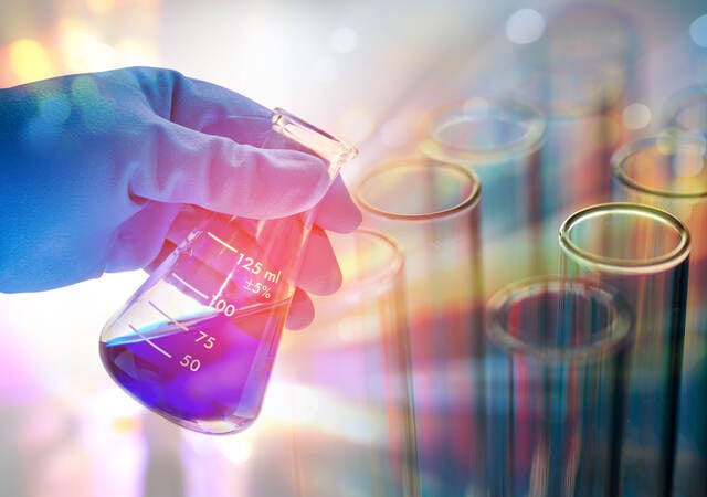 Rubber-gloved hand holding beaker containing blue liquid on a colorful background with test tubes