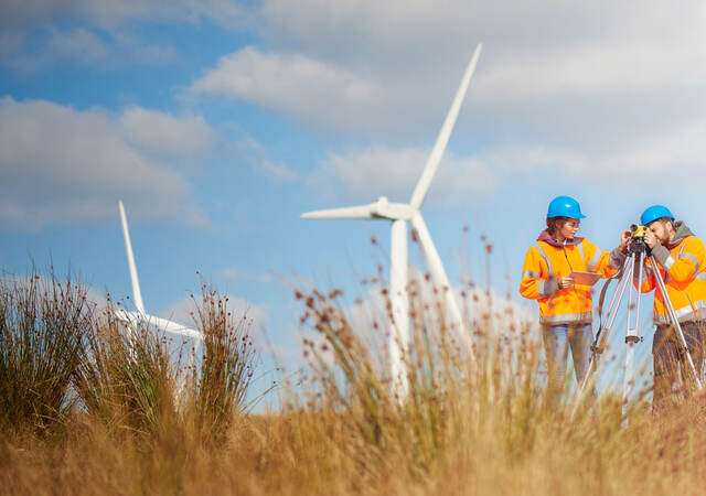 Two workers surveying in a field with wind turbines behind them