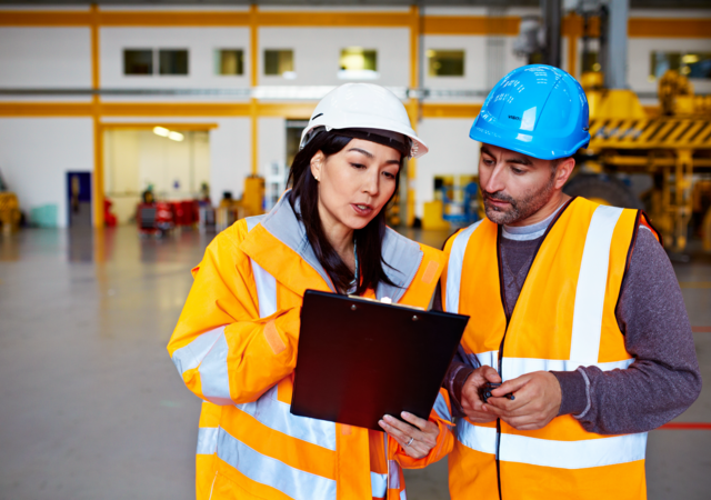 Two workers talking together over a clipboard while standing inside a large warehouse