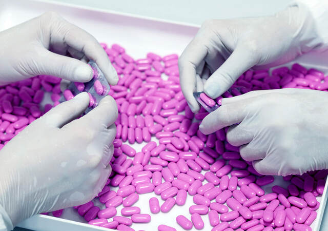 Two sets of hands in latex gloves remove purple pills from foil-backed medicinal packaging