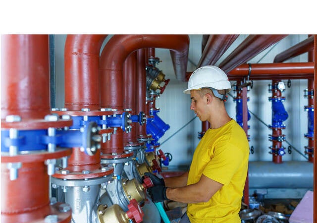 A male heating plant worker wearing a hardhat is standing next to a pipe system and performing a quality check.