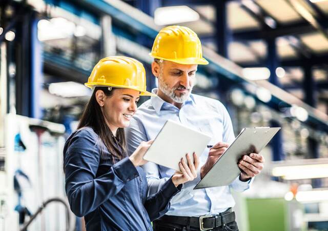 Two workers in yellow hardhats comparing notes on their tablet and clipboard 