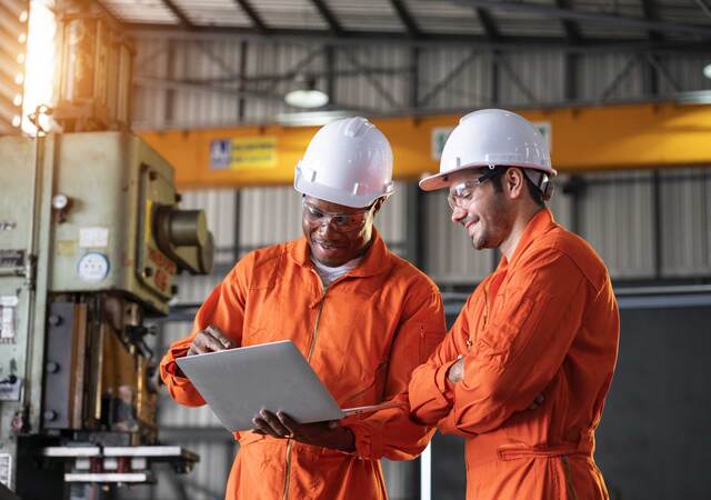 Two engineers in orange coveralls and white hardhats looking at a laptop