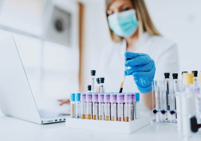 Close up of lab assistant holding test tube while wearing a lab coat, mask and rubber gloves