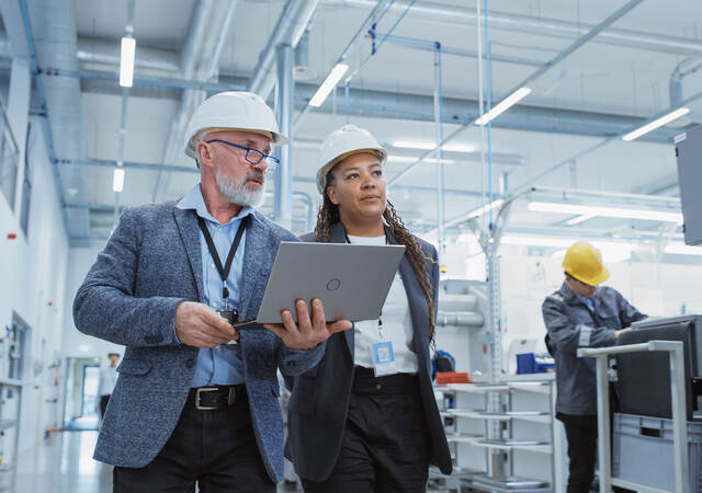 Two managers touring a factory floor while reviewing a laptop