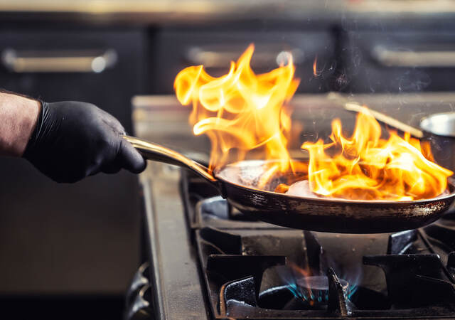 A chef wearing a protective glove while holding a frying pan that has flames coming off of it