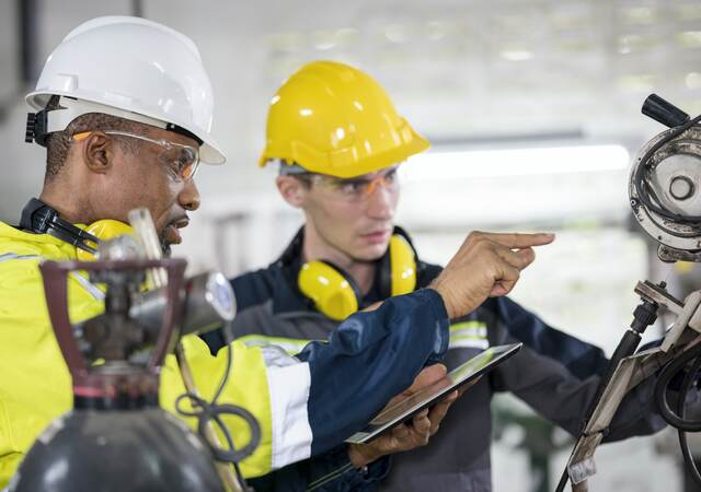 Factory line workers in safety gear examine and discuss a machine part.