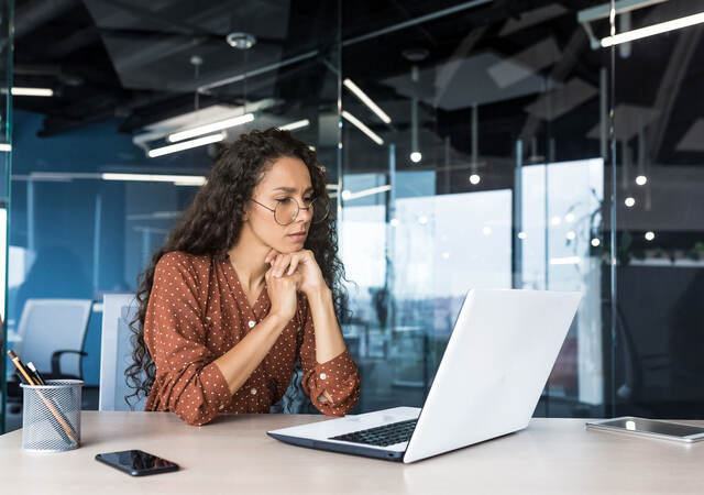 Person in a glass office, looking thoughtfully at a laptop