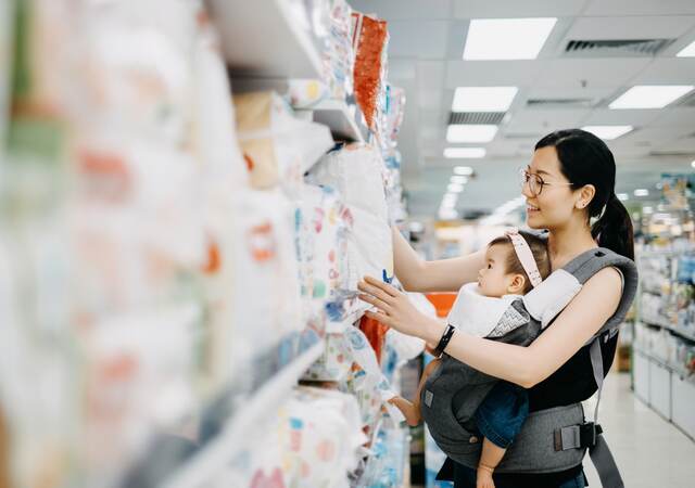 Young Asian mother carrying cute baby girl shopping for baby product in a shopping mall and is looking at a variety of diapers.