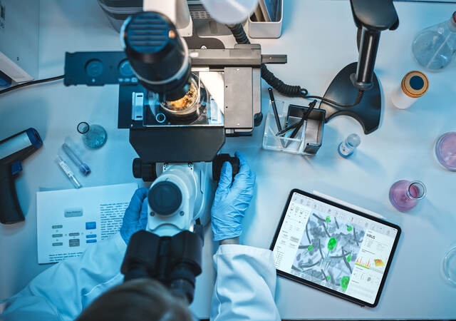 Top down view of a medical research scientist in blue rubber gloves using a microscope next to a tablet with virus data displayed