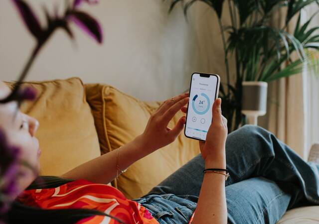 Relaxed woman reclines on a sofa and uses a thermostat app on her smart phone to control the housing heating system