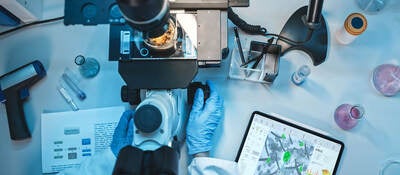 Top down view of a medical research scientist in blue rubber gloves using a microscope next to a tablet with virus data displayed