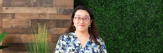 Vicky Zhang in floral shirt sits at table in front of vine covered wall. 