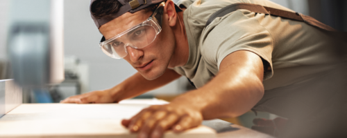 man with safety glasses lining a sheet of wood up for cutting on a table saw